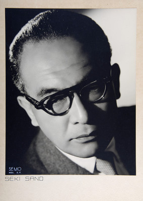 "Seki Sano", de frente con traje formal y anteojos, fondo obscuro, retrato