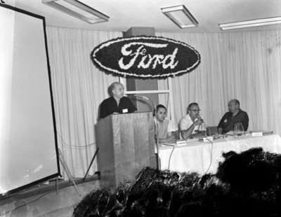 Hombre de mesa directiva con micrófono en mano durante discurso en la convención "ford" en un salón