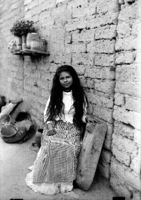 Niña con metate junto a muro de adobe, retrato