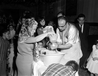 Sacerdote y mujer sostienen a niño en una tina, durante ceremonia de bautizo