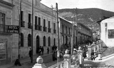 Gente deambula por una calle con comercios y hoteles, tarjeta postal